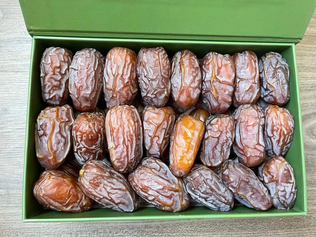 Kurma Medjool disusun tiga baris rapat dalam kotak hadiah hijau yang terbuka di atas meja kayu